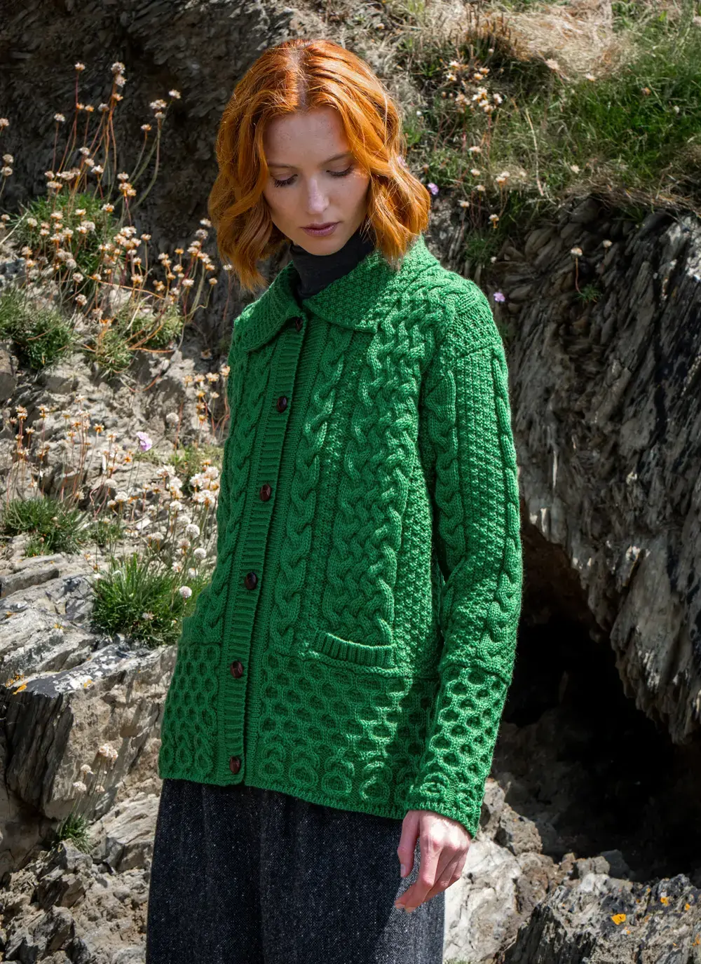Red head model wearing emerald green jackie aran lumber cardigan at bottom of cliff face